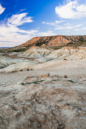 This captivating landscape highlights the rugged beauty of badlands terrain under a sunset sky. The foreground features textured earth leading to strikingly striped hills, with the sky above painted in soft streaks of white against a calming blue.