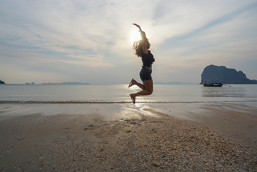 Happy Asian young woman in casual style fashion and straw hat jumping at sand beach. Relaxing, fun, and enjoy holiday at tropical paradise beach with sunset sky and white clouds.