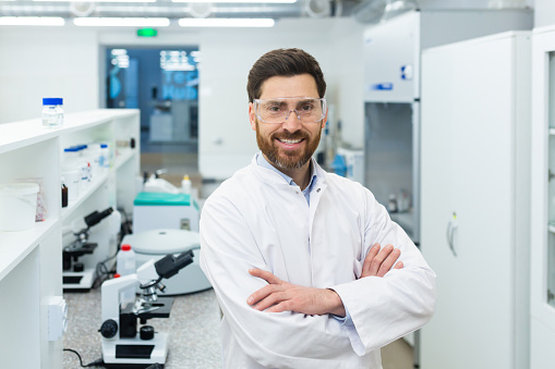 Portrait mature experienced laboratory assistant in a small laboratory, a man with a beard and glasses is smiling and looking at the camera among microscopes, a scientist in a white medical coat