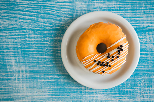 Bright orange donut decorated with glaze and chocolate balls on a blue wooden background, top view, copy space.
