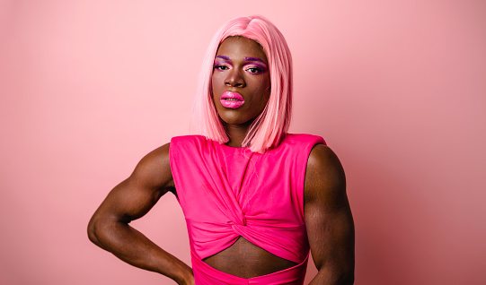 Young African American drag queen wearing pink wig and bright makeup standing with hand on waist in studio looking at camera