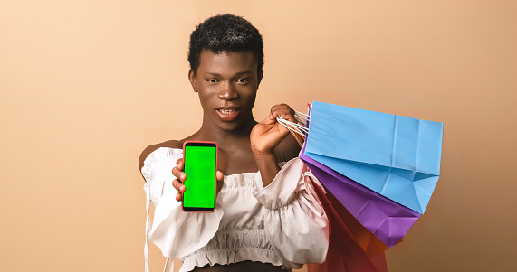 Confident African American transgender person holding colorful paper bags and showing smartphone with green screen to camera after successful online shopping against beige background