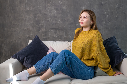 a young woman smiles dreamily while sitting on the sofa. casual comfortable clothes and gray wall background