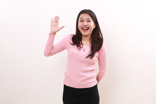 Cheerful excited young asian girl standing while raising her hand greeting gesture, isolated on white