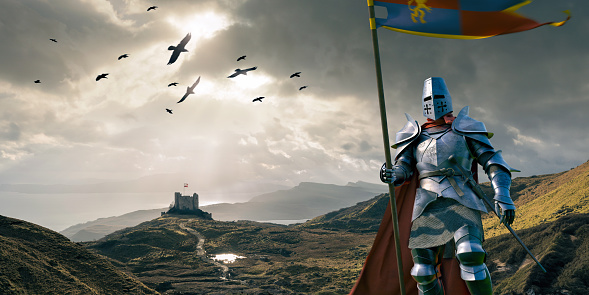 A close up image of a medieval knight dressed in full armour, with chainmail and red cape, holding a pole with a banner flag blowing in the wind. The warrior is standing on hilly coastal ground with a castle, and birds flying in the background. With motion blur.