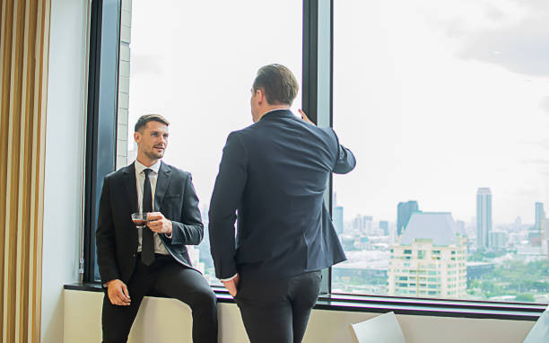 istock Two Caucasian smart businessmen wearing formal suits, talking with relaxation, taking break, drinking coffee, standing in modern indoor office beside window. Business Concept. 1806143060