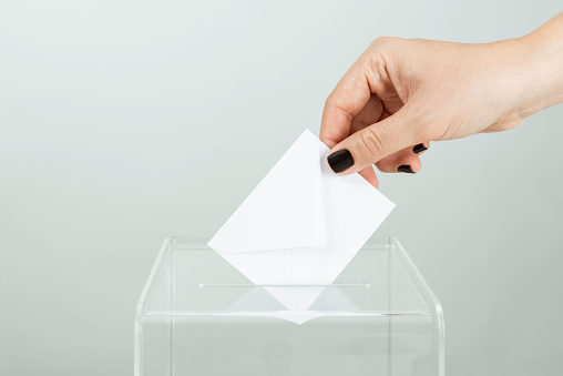 Human hand is inserting white envelope into ballot box.