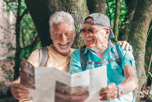 Cheerful couple of senior male friends sitting looking at footpath map in a trekking day in the forest enjoying nature and healthy lifestyle in vacation or retirement