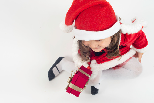 Little girl in santa claus costume is holding a gift box and is sitting on white background.