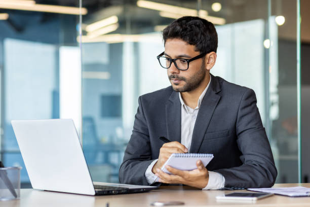 istock Serious and concentrated thinking businessman watching webinar online course, man upgrading at workplace inside office, hispanic man taking notes and writing data in notebook 1806217903