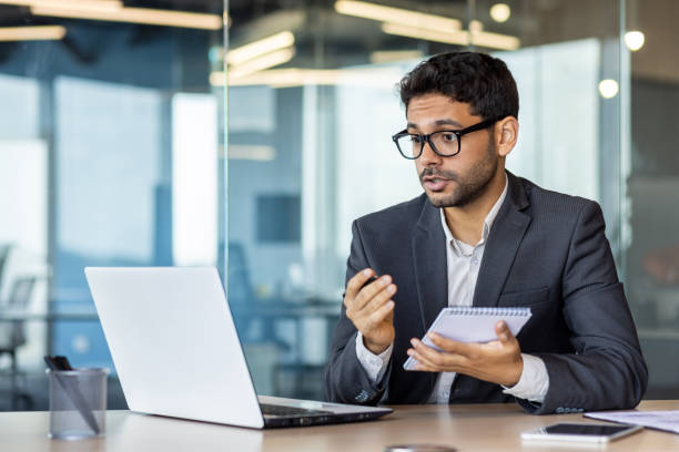 istock Young Indian male office worker sitting at a desk in the office and using a tablet, talking on a video call through a laptop 1806218426