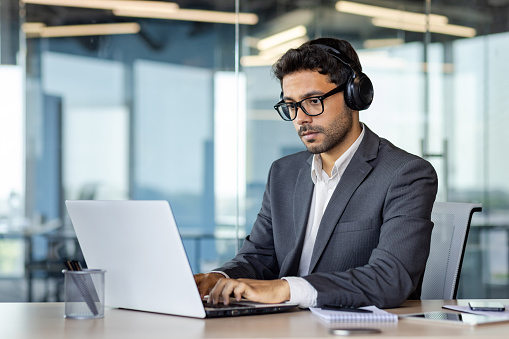 Serious man working inside office with laptop, concentrated and thinking businessman boss in business suit using headphones listening to music and educational audio course and book.