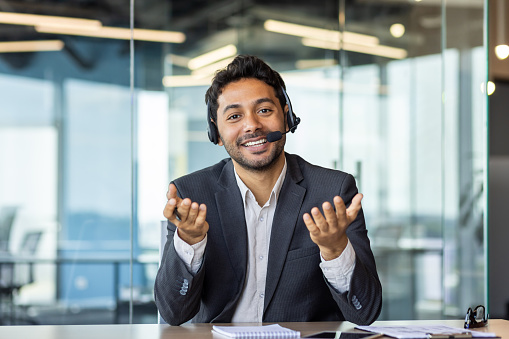 Portrait of a young Indian man who works in the office, sits at a desk in a headset, talks and consults remotely. Gesturing with his hands and looking at the camera with a smile