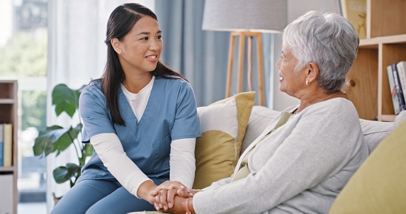 Home, nurse and old woman with conversation, holding hands or discussion with health, compassion or results. Apartment, caregiver or elderly person on a sofa, support or comfort with empathy or relax