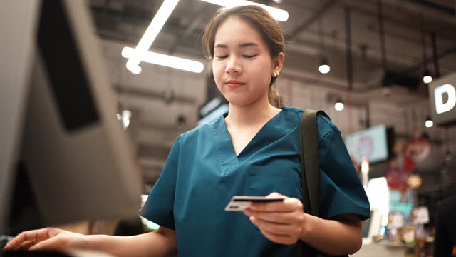 Young Asian female healthcare doctor in blue medical scrubs buying food and drink at supermarket, Woman paying credit card at shopping mall, Lifestyle in scrubs