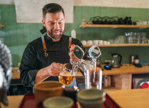 istock Smiling stylish bearded barman dressed black uniform with an apron tapping fresh lager beer into glass mug at bar counter. Successful people, beer consumption, beverages industry concept image 1806250402