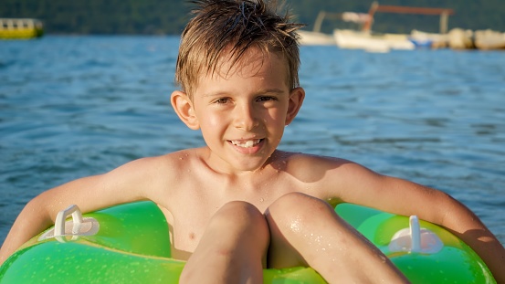 Happy smiling boy sitting in green inflatable ring and swimming in the sea towards camera. Family holidays, vacations, summertime