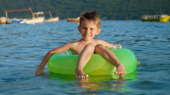 Happy cheerful boy splashing water while sitting in inflatable ring and swimming on calm sea waves at sunset. Family holidays, vacations, summertime