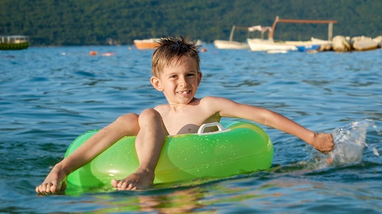 Happy cheerful boy splashing water while sitting in inflatable ring and swimming on calm sea waves at sunset. Family holidays, vacations, summertime