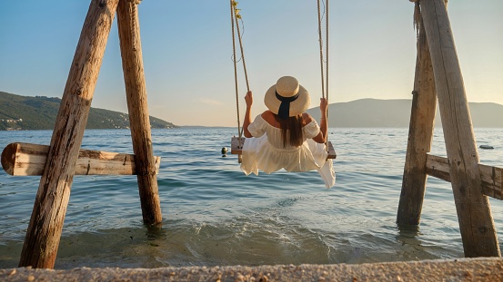 Rear view of young elegant woman in dress relaxing on beach at sunset and swinging on swing. Tourism, summertime, holiday and vacation on beach