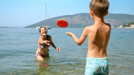 little boy throwing frisbee to his mother standing in the sea next to the beach. Family holiday, vacation and fun summertime of children and parents