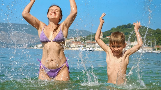 Happy cheerful excited boy splashing water with mother while relaxing on sunny sea beach.