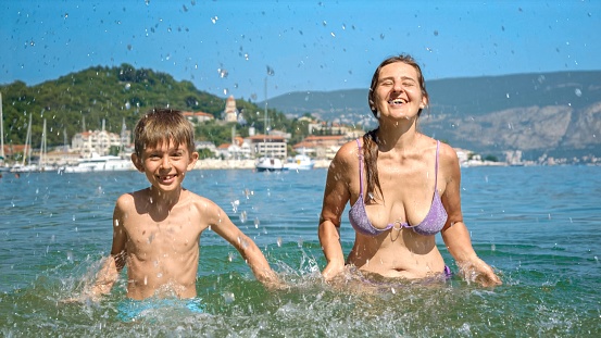 Mother and her son playing and splashing water at the sea beach in slow motion. Family holiday, vacation memories, and the pure joy of summertime.