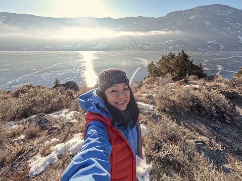 A solo senior Chinese woman takes a selfie for social media while winter hiking to a snowy viewpoint of an ice covered lake at sunrise.  Merritt, British Columbia, Canada.