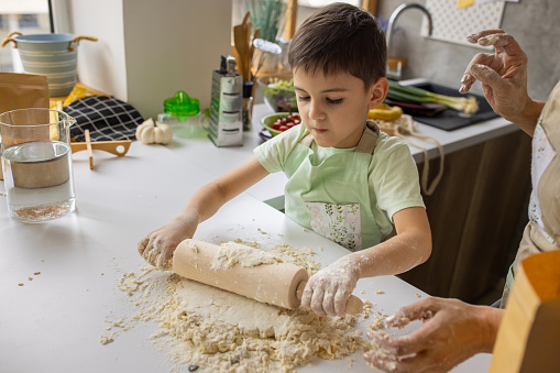 Little boy rolling dough with rolling pin, while his grandmother is helping him in kitchen at home