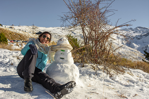 latin woman sitting on frozen ground next to snowman,