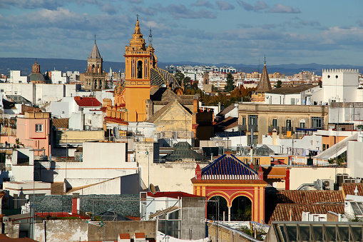 Photo with the view of part of the city of Seville (Andalusia region, southern Spain) with a yellow church in the center of the photo and numerous bell towers around it against a background of a blue sky with clouds
