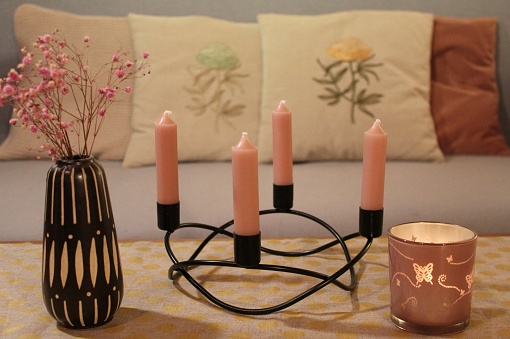 Modern Advent wreath made of black metal, pink candles, vase with dried flowers. A sofa with embroidered cushions in the background.