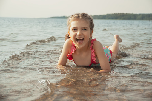 Happy laughing child girl of 5 years in wet blue swimsuit lies on shore in sea water on sunny summer day, lifestyle, copy space