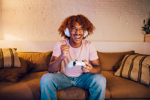 Young man with white headphones playing video games on a sofa in a living room. Happy young man celebrating a winning