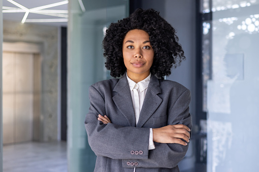 Portrait of serious successful African American business woman, boss in business suit looking focused at camera with crossed arms, female financial worker inside office at workplace.