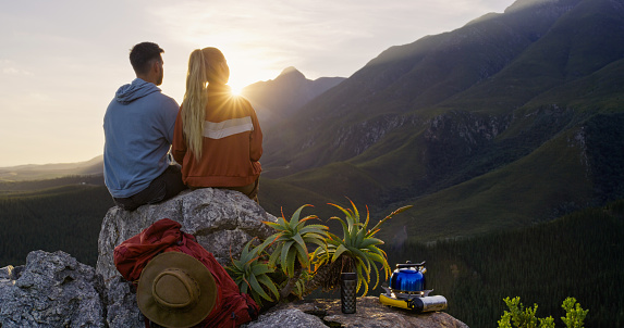Couple, sunrise or hiking on mountain in nature for travel, adventure or experience with peace and freedom. Rear view, man or woman sitting on cliff for scenery, outdoor or relax on hill for vacation