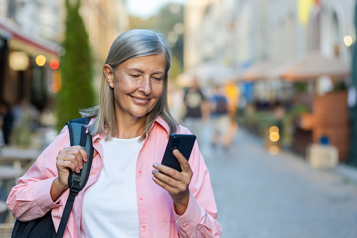 Mature female tourist smiling typing a message on the phone, traveling woman retired gray-haired pensioner using smartphone app while walking in the evening city smiling contentedly.