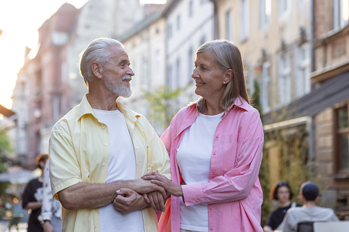 Senior couple of gray-haired man and woman standing holding hands on a city street and smilingly looking at each other.