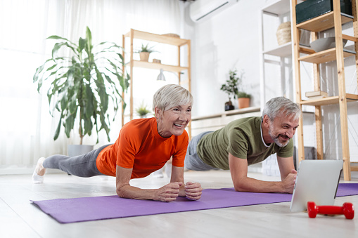 A wife and a husband attend an online fitness class in their apartment. They are doing plank and looking at the digital tablet.