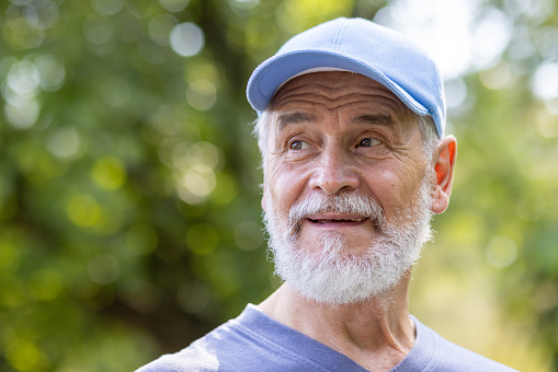 Close-up portrait of a gray-haired senior man with a beard in a cap doing sports, walking in the park outdoors. He looks away with a smile.