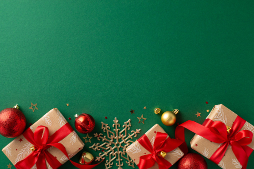 Festive preparations underway! Top view of craft paper gift boxes, red and gold baubles, snowflake decor on green background. Confetti bring holiday cheer. Personalize the space with your message