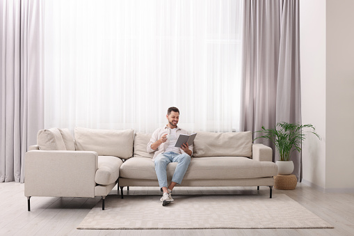Happy man reading book on sofa near window with beautiful curtains in living room