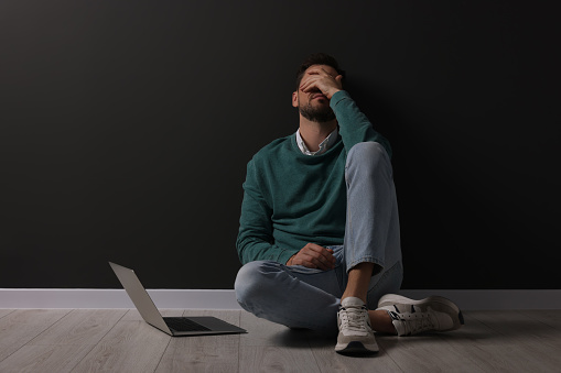 Upset man sitting on floor near laptop against black wall. Space for text
