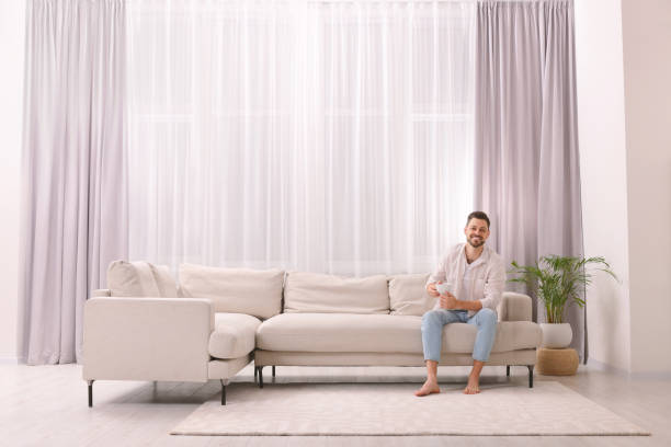 istock Happy man drinking coffee while resting on sofa near window with beautiful curtains in living room 1806471988