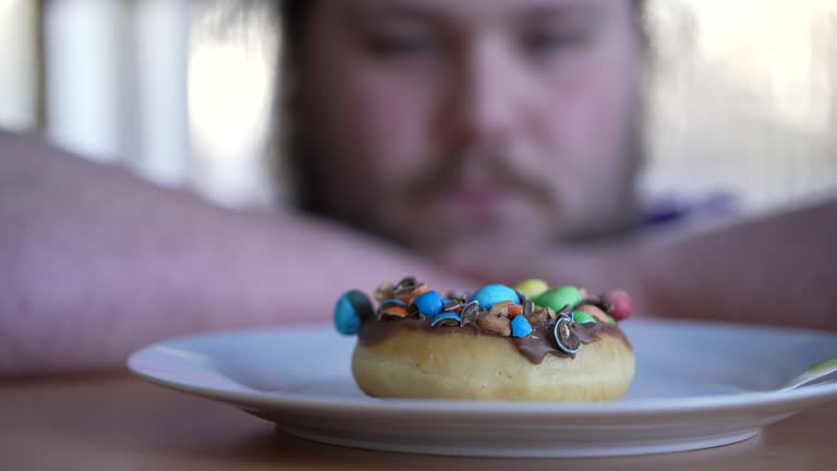 Overweight Person wanting to eat sugar food on plate. One Young large build man in diet fasting from junk food. Donut closeup in front of large build guy. Weight loss concept
