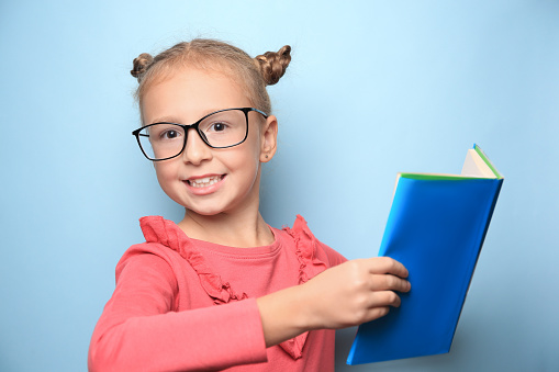 Cute little girl with glasses and textbook on light blue background