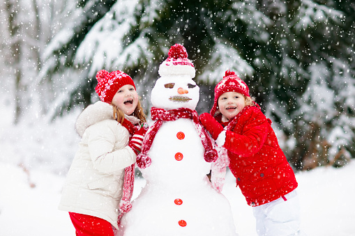 Children build snowman. Kids building snow man playing outdoors on sunny snowy winter day. Outdoor family fun on Christmas vacation. Boy and girl play snow balls. Winter clothing for baby and toddler.