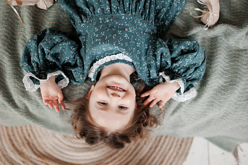 Child birthday celebration. Top view of happy little girl 4-5 years wears stylish rustic style dress is lying on bed, smiling and looking at camera