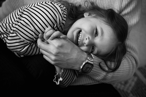 Happy children's day. Black and white photo of unrecognisable dad with adorable laughing little daughter are having fun spending time together on weekend at home. Playful child girl and daddy hugging.