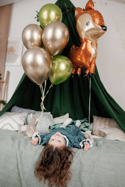 comemoração de aniversário da criança. vista superior da menina feliz 4-5 anos usa vestido estilo rústico elegante está deitado na cama, sorrindo e olhando para a câmera. quarto decorado para festa de aniversário com balões - preschool 2 3 years preschooler little girls - fotografias e filmes do acervo